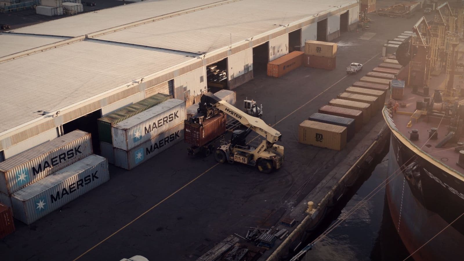 A container handler unloading containers beside a warehouse dock at sunrise